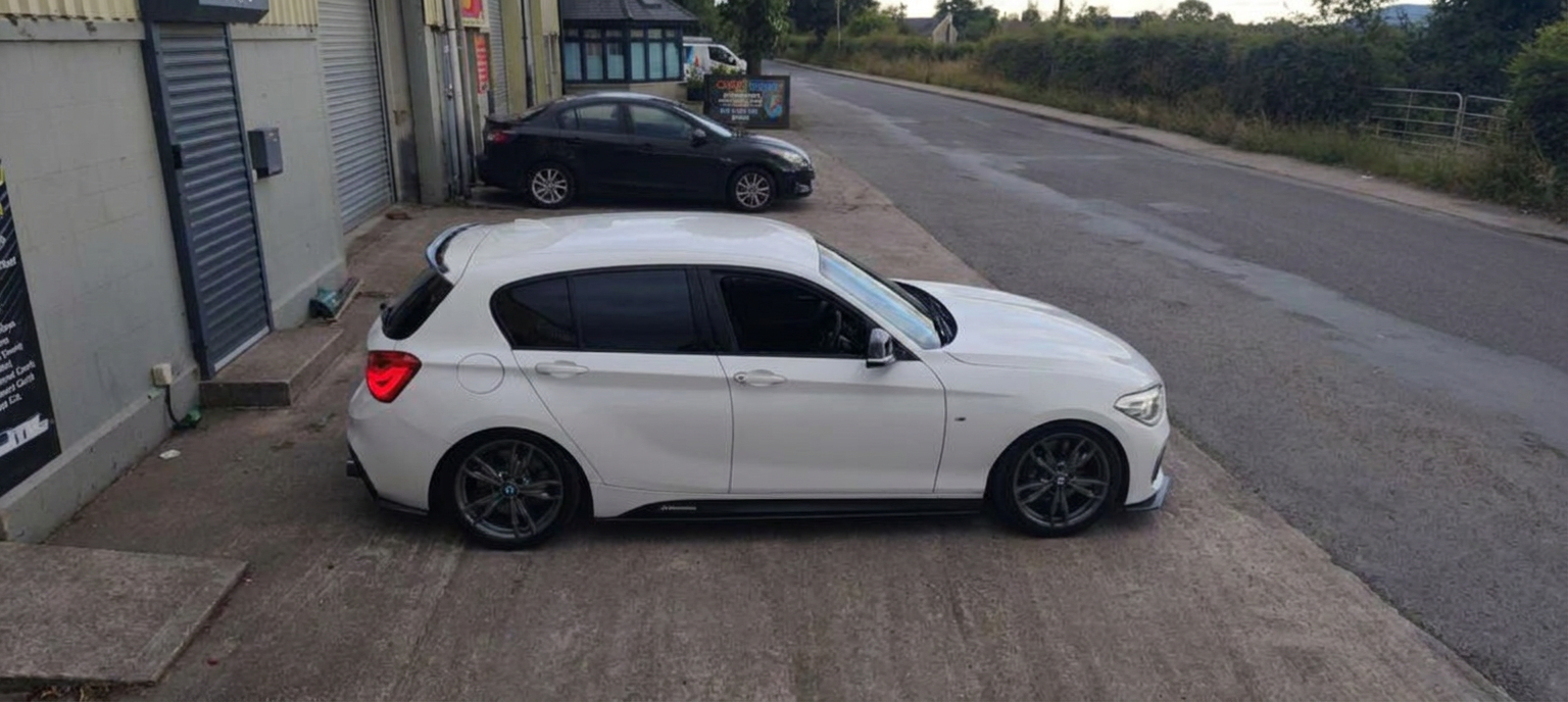 High three-quarter side view of a tinted white BMW 1 Series hatchback on the workshop forecourt